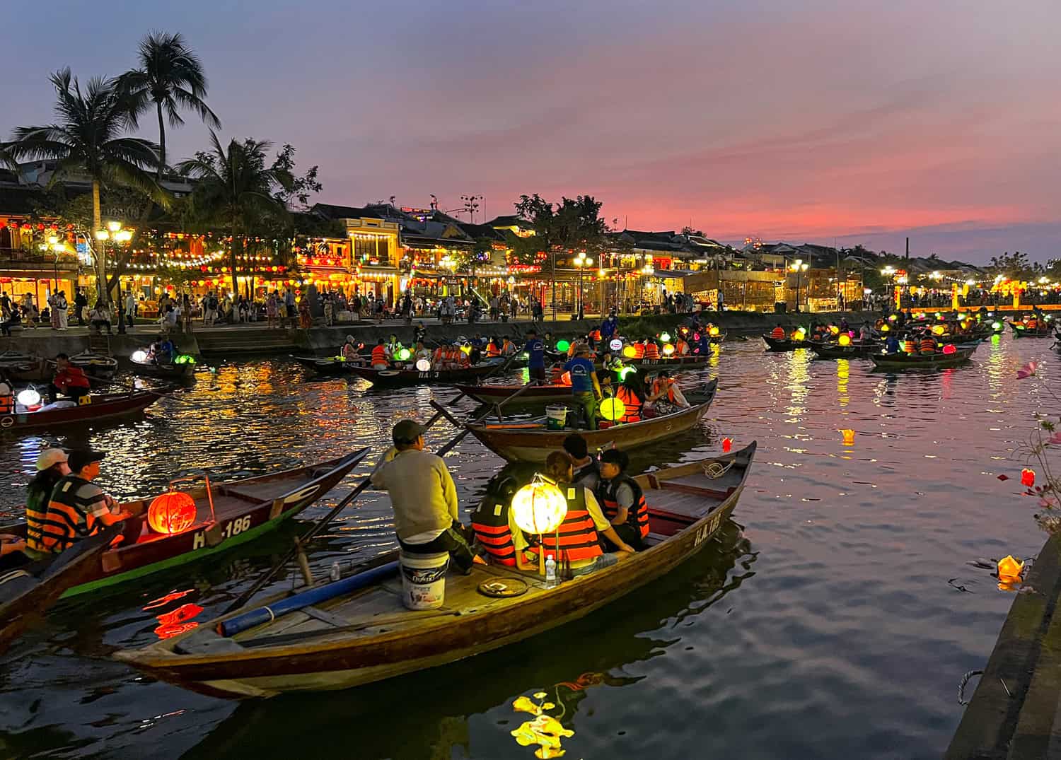 Một chiếc thuyền gỗ nhỏ với đèn lồng trôi trên sông Thu Bồn vào ban đêm ở Hội An, với các tòa nhà được chiếu sáng ở hậu cảnh.